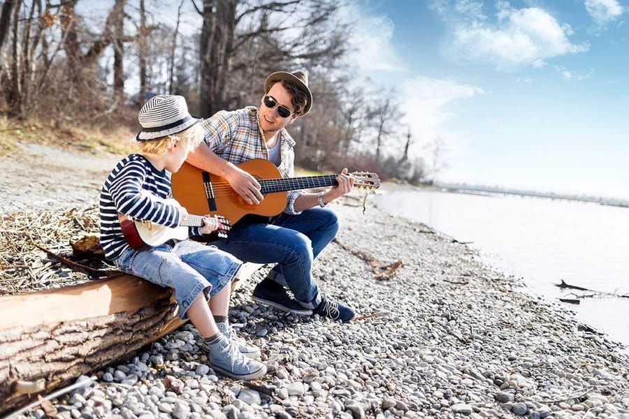 Vater und Sohn sitzen am Ufer eines Flusses, beide spielen auf einer Gitarre, das Bild ist Teil der Familienkampagne "Ich bin Vorbild"